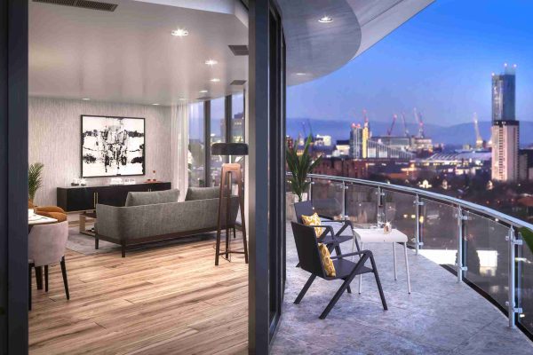 View from inside the living room looking out onto a private balcony equipped with outdoor seating. The background features a clear view of the Manchester cityscape and nearby MediaCityUK lights in the evening.